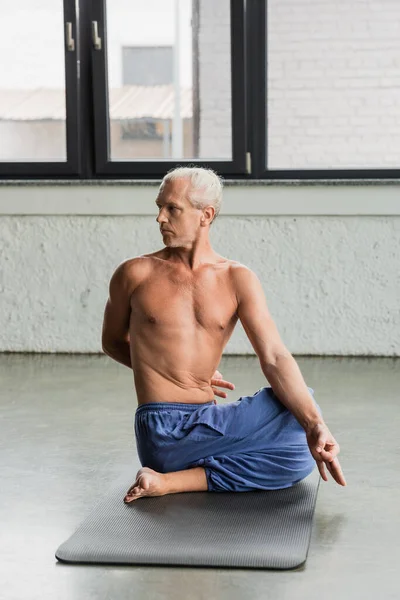 Shirtless and grey haired man in pants sitting in half lord of fishes pose and doing gyan mudra - foto de stock