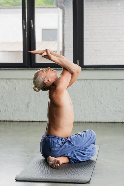 Uomo senza maglietta e dai capelli grigi in pantaloni seduto in posa yoga con le mani attorcigliate — Foto stock