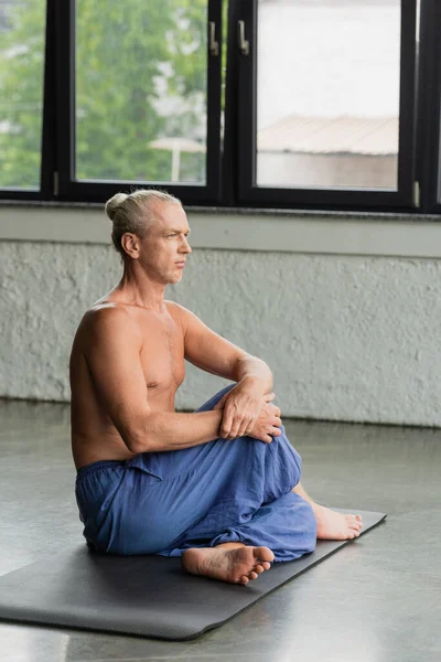 Босоніж і сіра волохата людина в штанях, сидячи зі скрученими ногами на йога килимок, роздумуючи в студії — стокове фото