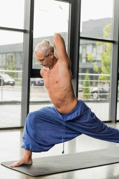 Homme aux cheveux gris en pantalon bleu faisant pose croissant sur tapis de yoga — Photo de stock