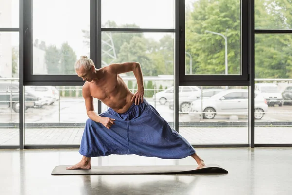 Shirtless and grey haired man in pants doing extended side angle pose in yoga studio — Stock Photo