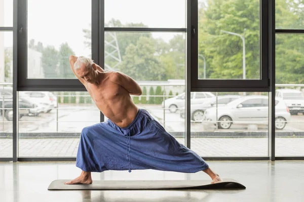 Hombre de pelo gris haciendo pose de ángulo lateral extendido en estudio de yoga - foto de stock