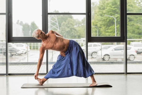 Grey haired man practicing yoga triangle pose on mat in studio — Fotografia de Stock