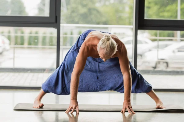 Grey haired man doing wide legged forward bend on yoga mat in studio — Photo de stock