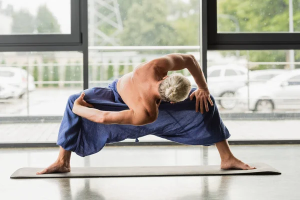 Grey haired man doing wide legged forward fold twist while practicing yoga — Fotografia de Stock