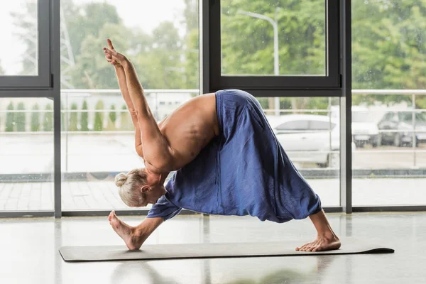 Вид сбоку седого волосатого человека, делающего позу пирамиды с поднятыми руками на коврике для йоги в студии — стоковое фото