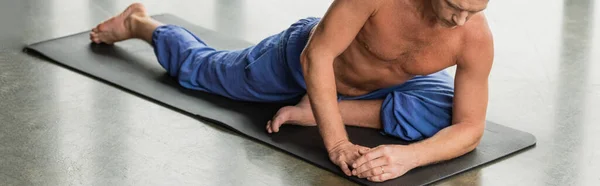 Shirtless man doing half pigeon pose on mat in yoga studio, banner - foto de stock