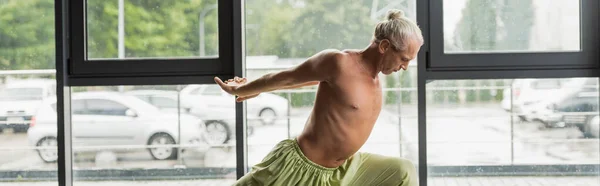 Grey haired man in green pants practicing crescent lunge in yoga studio, banner - foto de stock