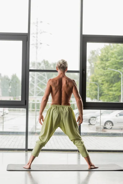 Back view of shirtless man in green pants meditating on yoga mat in studio — Stock Photo