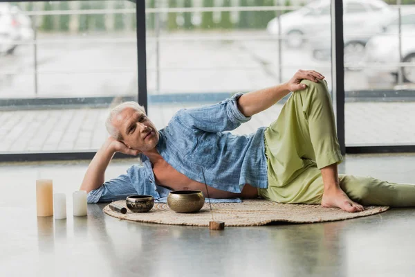 Grey haired man resting near Tibetan singing bowls and candles in yoga studio — Stock Photo