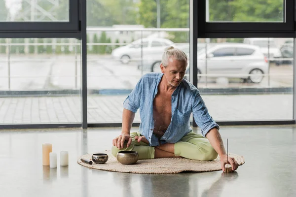Grey haired man moving incense stick near Tibetan singing bowls and candles in yoga studio - foto de stock
