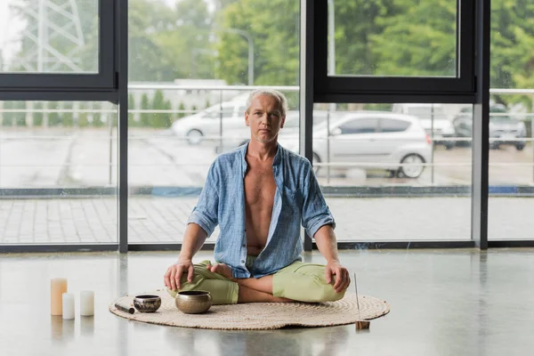 Grey haired man meditating near Tibetan singing bowls and incense stick in yoga studio — Photo de stock