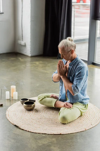Grey haired man meditating with praying hands near Tibetan singing bowls and incense stick in yoga studio — Photo de stock