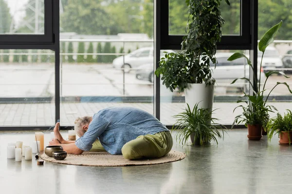 Grey haired man stretching back near Tibetan singing bowls and candles in yoga studio — Stock Photo