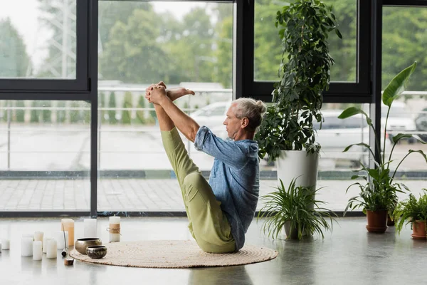Grey haired man doing boat yoga pose near Tibetan singing bowls and incense stick in yoga studio — Stock Photo