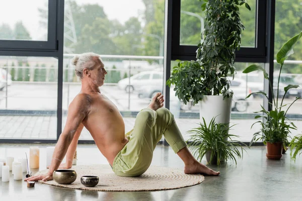 Side view of shirtless man stretching back near candles and Tibetan singing bowls in yoga studio — стокове фото