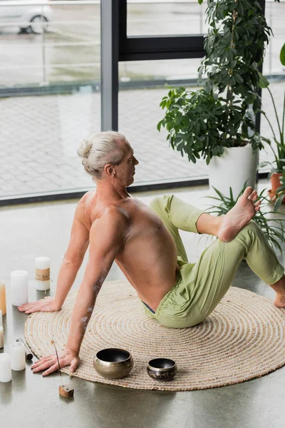 Side view of grey haired man stretching back in yoga pose near candles and Tibetan singing bowls in studio — Stock Photo