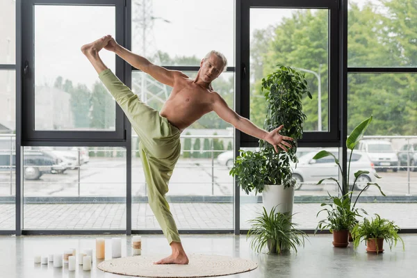 Uomo senza maglietta in pantaloni verdi facendo mano alla posa alluce vicino a candele e piante in studio di yoga — Foto stock