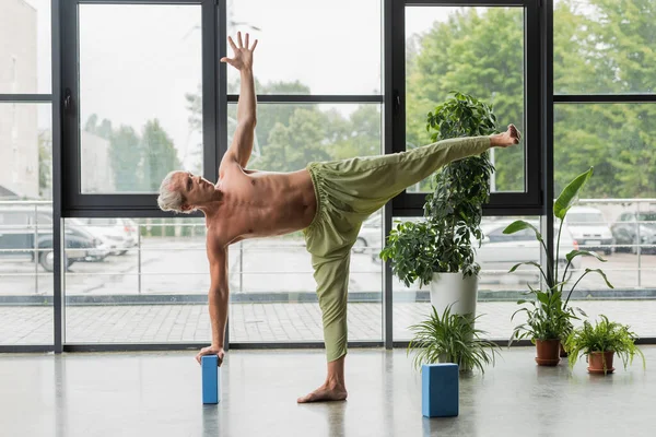 Homem sem camisa em calças verdes fazendo meia lua pose perto de bloco de espuma de ioga em estúdio — Fotografia de Stock