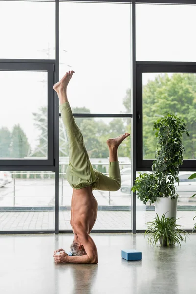Hombre de pelo gris haciendo soporte de cabeza apoyado cerca de bloque de espuma de yoga - foto de stock