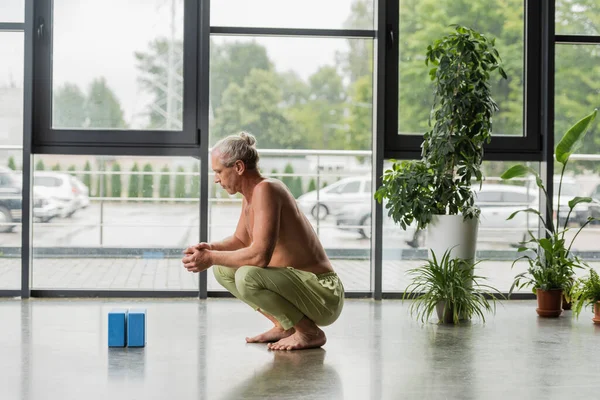 Side view of shirtless and grey haired man sitting with clenched hands and looking at blue yoga foam blocks in studio — Stock Photo