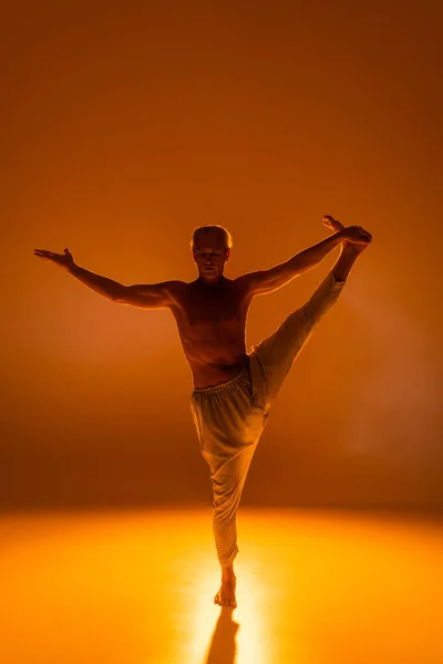 Full length of shirtless man in pants doing hand to big toe pose on orange background - foto de stock