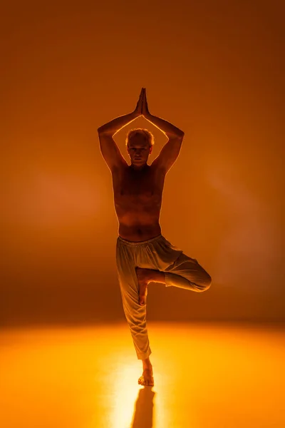 Full length of shirtless man in pants standing in tree pose on orange background — Stock Photo