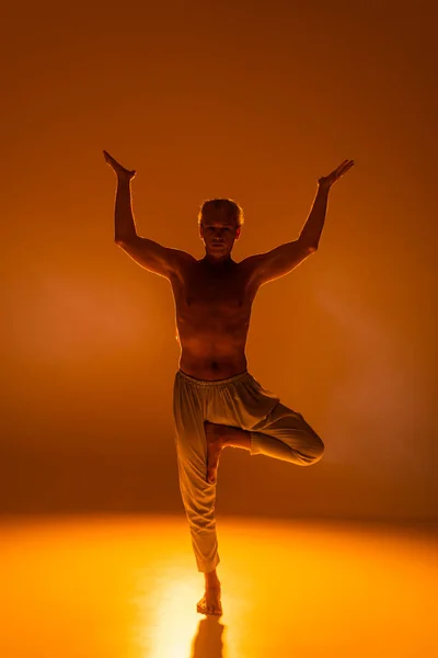 Full length of shirtless man in pants standing in tree pose on orange background — Stock Photo