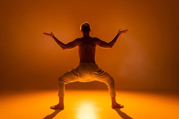 Вид сзади безрубашечника в штанах, практикующего йогу, позирующего на оранжевом фоне — стоковое фото