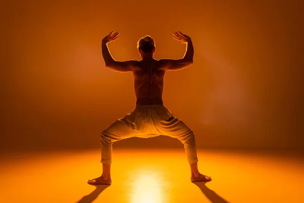 Back view of shirtless man practicing goddess yoga pose on orange background — Stock Photo