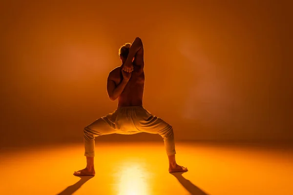 Вид сзади дерзкого человека, практикующего йогу в позе богини со сжатыми руками за спиной на оранжевом фоне — стоковое фото