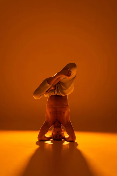 Longitud completa de hombre sin camisa haciendo postura de cabeza de yoga en marrón - foto de stock