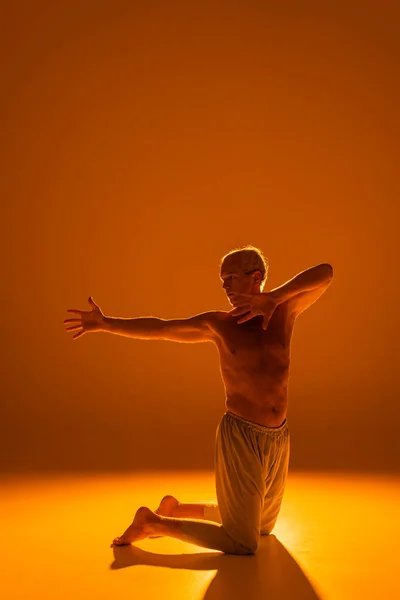 Full length of shirtless man standing on knees while meditating on brown background — Stock Photo