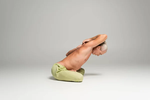 Side view of shirtless man sitting in lotus pose with clenched hands behind back on grey background — Stockfoto