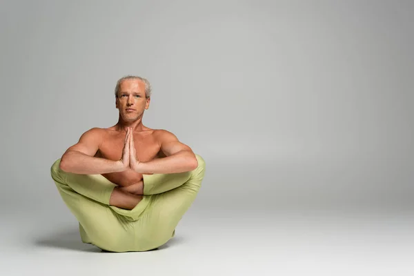 Shirtless man in pants sitting in womb embryo pose and doing anjali mudra on grey — Stock Photo
