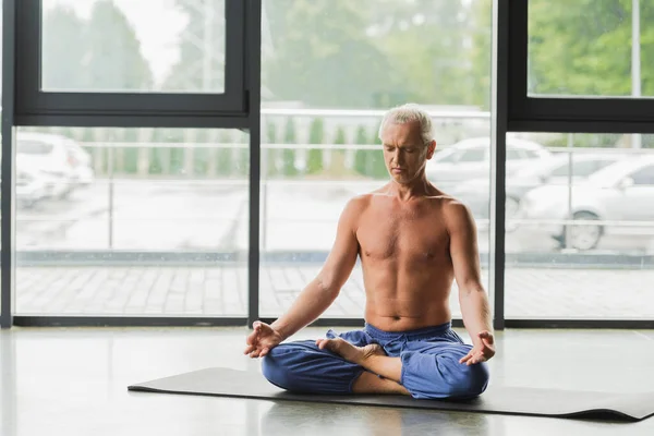 Hombre descalzo en pantalones azules sentado en pose de loto y haciendo gyan mudra en estudio de yoga - foto de stock