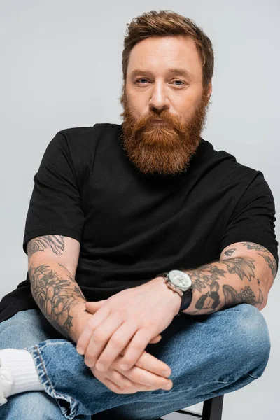 Tattooed bearded man in black t-shirt and jeans sitting and looking at camera isolated on grey — Stockfoto