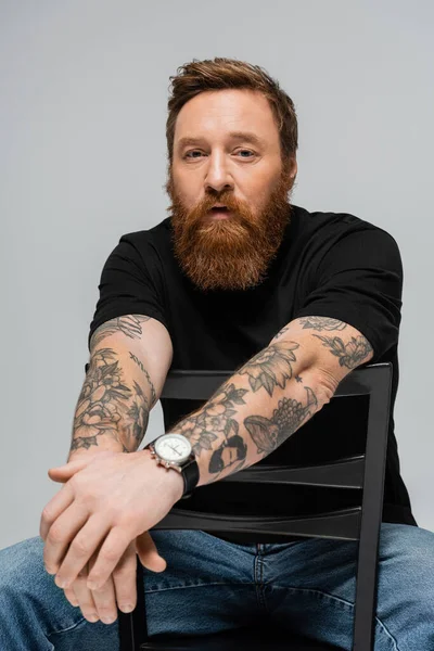 Вдумливий бородатий чоловік в чорній футболці і наручний годинник, сидячи на стільці і дивлячись на камеру ізольовано на сірому — стокове фото