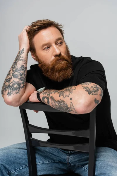 Dreamy bearded man in black t-shirt looking away while sitting on chair isolated on grey - foto de stock