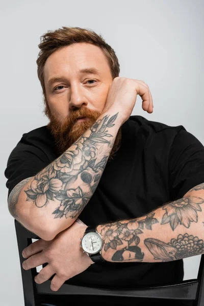 Стомлений татуйований чоловік в чорній футболці і наручних годинниках сидить на стільці і дивиться на камеру ізольовано на сірому — стокове фото