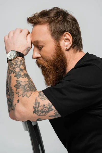 Side view of tired tattooed man in black t-shirt and wristwatch sitting with closed eyes isolated on grey — Stock Photo