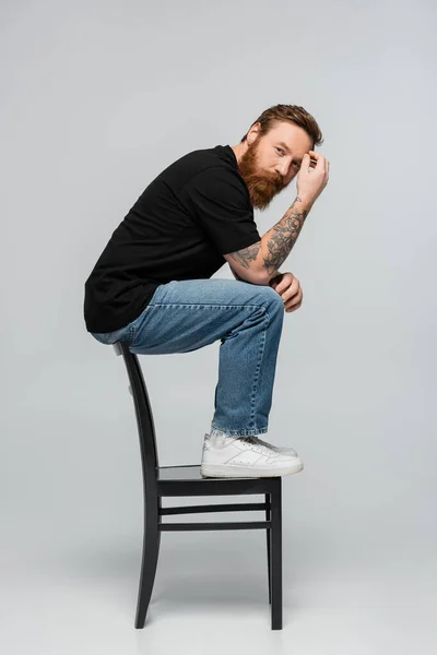 Full length of thoughtful bearded man in black t-shirt and jeans sitting on chair back and looking at camera on grey background — Stockfoto