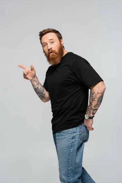 Surprised bearded man in black t-shirt and jeans looking at camera and pointing with finger isolated on grey - foto de stock