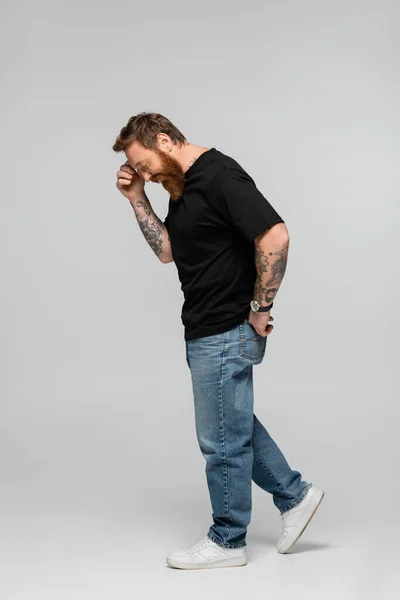 Side view of smiling man in black t-shirt and jeans touching forehead while smiling on grey background — Stockfoto