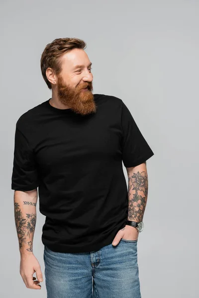 Весёлый бородатый мужчина в черной футболке, стоящий с рукой в кармане джинсов и смотрящий в сторону изолированный на сером — стоковое фото
