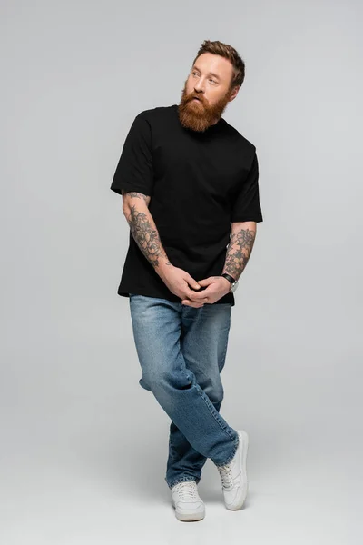 Повна довжина татуйованого бородатого чоловіка в джинсах і чорній футболці, що стоїть з зіпсованими руками і дивиться на сірий фон — стокове фото