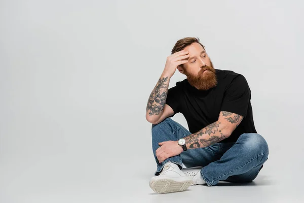 Longitud completa de hombre tatuado reflexivo en camiseta negra y jeans mirando hacia otro lado mientras está sentado sobre fondo gris - foto de stock