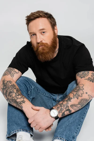 Serious bearded man in black t-shirt and jeans looking at camera while sitting on grey background — Photo de stock