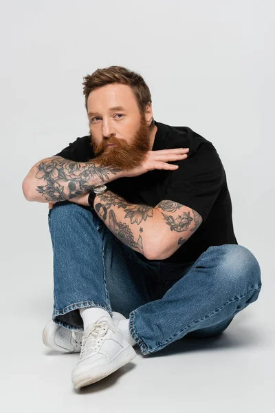 Elegante uomo tatuato in blue jeans e sneakers bianche seduto con le gambe incrociate e guardando la fotocamera su sfondo grigio — Foto stock