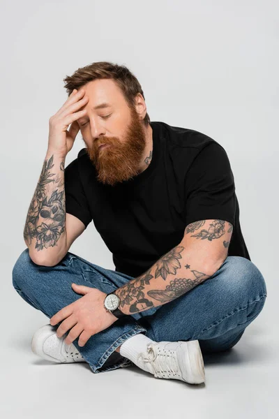 Tired bearded man in jeans sitting with crossed legs and hand near head on grey background — Stock Photo